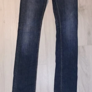 Mörkblå Nudie Jeans W26 L34 - Snygga mörkblå jeans från Nudie Jeans med klassisk orange söm och vågig brodyr på bakfickan. Modellen har rak passform och normal midja, tillverkade i slitstarkt bomullsmaterial. Perfekt för dig som gillar stilrena och tidlösa jeans.. Slim fit modell 