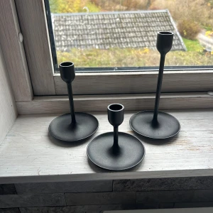Ljusstake  - Bilderna visar tre svarta ljusstakar i metall med modern, minimalistisk design. De har olika höjder och rund fot, vilket ger ett stilrent intryck.aldrig använda 