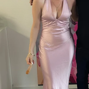 Ljuslila långklänning i satin - Säljer en elegant långklänning i ljuslila satin med djup v-ringning. Klänningen har en figurnära passform och en mjukt fallande kjol. Perfekt för dig som vill sticka ut med en stilren och glansig look. Djup urringning på ryggen. Använd 1 gång. Perfekt för fest/bal. Inköpt på NewYorker för 399kr. 