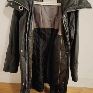 Svart lång parkajacka med dragkedjor - Svart lång parkajacka från Etnic med quiltad insida och flera praktiska fickor med dragkedjor. Jackan har huva, detaljer med metallöljetter och justerbara ärmslut. Perfekt för kalla dagar och har en snygg urban look.