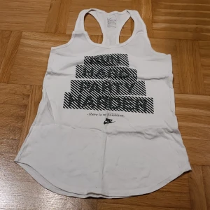 Vit linnetopp från Nike XS - Säljer en vit linnetopp från Nike i storlek XS. Toppen har ett svart grafiskt tryck med texten 'Run Hard Party Harder' och Nike-logga framtill. Modellen är ärmlös med brottarrygg och tillverkad i mjuk bomull, perfekt för träning eller chill.