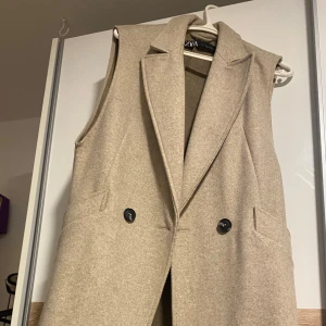 Beige lång väst från ZARA - Stilren lång beige väst från ZARA med klassisk krage och två svarta knappar framtill. Västen har fickor på sidorna och är tillverkad i ett mjukt ull-liknande material. Perfekt för lager-på-lager och ger en clean look. Helt ny, fick ingen användning av den därför väljer jag att sälja den.