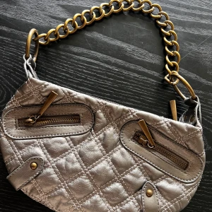 Quiltad  silver axelväska med kedja - Snygg quiltad axelväska i metallic silver med grov guldfärgad kedja som axelrem. Väskan har två dragkedjefickor framtill och detaljer i mässing. Tillverkad i skinnimitation och har ett ljust innerfoder. Perfekt storlek för det viktigaste.