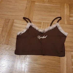 Brun ribbad linnetopp med spets - Säljer en brun croppad linnetopp med texten Spoiled med tunna axelband och ljus spetskant med blommönster. Toppen har ribbad struktur och texten 'Spoiled' broderad framtill. Perfekt för dig som gillar söta detaljer och skön passform.