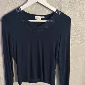 Mörkblå långärmad topp med spets - Snygg mörkblå långärmad topp från Gina Tricot med v-ringad hals och dekorativ spetsdetalj runt ringningen. Tunn och mjuk kvalitet som sitter skönt mot kroppen. Perfekt att styla med jeans eller kjol för en clean look.