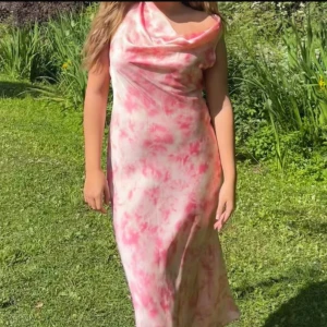 Rosa och vit långklänning i satin - Supersnygg långklänning i satin med rosa och vitt mönster. Klänningen har en draperad urringning, smala axelband och öppen rygg med knyt. Sval och flowy passform, perfekt för varma dagar eller speciella tillfällen.