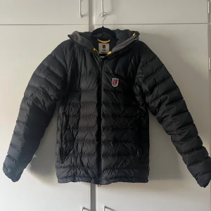 Svart Fjällräven Expedition dunjacka XS - Svart dunjacka från Fjällräven Expedition Series med gul snörning i huvan och klassisk röd logotyp på bröstet. Jackan har quiltad design, dragkedja framtill och två sidofickor. Perfekt för kalla dagar och har en snygg, sportig look.
