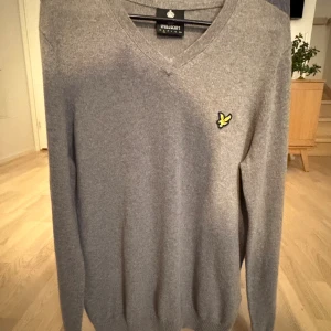 Grå v-ringad tröja från Lyle & Scott - Grå v-ringad tröja från Lyle & Scott med klassisk gul fågel-logga på bröstet. Tröjan är långärmad och har ribbade muddar vid ärmslut och nederkant. Materialet är mjukt och stickat, perfekt för dig som gillar stilrena och enkla plagg.