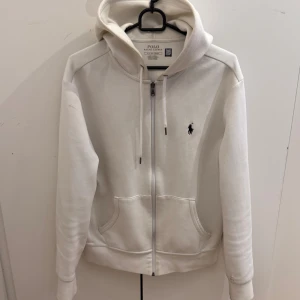 Polo Ralph Lauren zip-up hoodie - Vit hoodie från Polo Ralph Lauren med dragkedja och huva. Klassisk design med broderad logga på bröstet, två fickor framtill och snörning i huvan. Tillverkad i mjuk bomull för skön komfort. Perfekt för chill dagar och enkel att matcha med jeans eller joggers.
