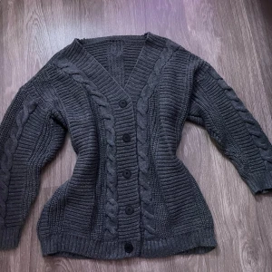 Mörkgrå stickad kofta med knappar - Säljer en mörkgrå stickad kofta med grova flätstickade detaljer längs ärmar och framstycke. Koftan har v-ringad hals och stora svarta knappar framtill. Perfekt för dig som gillar mysiga och stilrena plagg med lite extra struktur.