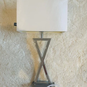 Lampa - Modern bordslampa med stilren, kromad metallfot i X-form och vit fyrkantig lampskärm. Lampan är i mycket gott skick utan synliga defekter eller slitage. Perfekt för sovrum, vardagsrum eller kontor och ger ett elegant intryck till inredningen. Strömkabel medföljer.