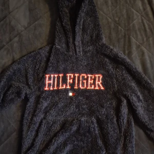 Mörkblå fluffig hoodie Tommy Hilfiger - Supermjuk och fluffig mörkgrå hoodie från Tommy Hilfiger med stor rödvit broderad logga på bröstet. Tröjan har huva och långa ärmar, perfekt för kyliga dagar. Materialet är fleece och ger en mysig känsla. Snygg och bekväm att dra på sig när man vill chilla.