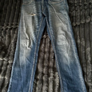 Blå raka jeans från Tiger of Sweden - Säljer ett par klassiska blå jeans från Tiger of Sweden med raka ben och snyggt slitna detaljer. Jeansen har fem fickor, normal passform och är tillverkade i robust denim. Perfekt för dig som gillar en avslappnad stil.