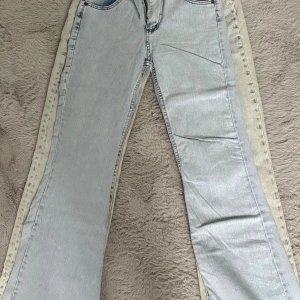 Bootcut ljusa jeans med kontrastkant - Snygga ljusblå jeans med bootcut passform och kontrastfärgade kanter längs benen. Klassisk femficksmodell med dragkedja och knapp framtill. Perfekta för dig som gillar retrovibbar och vill sticka ut lite extra.