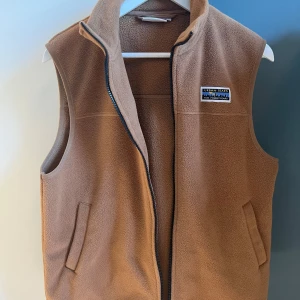Brun fleeceväst  - Säljer en brun fleeceväst från yunkyard med dragkedja och en liten logotyp på bröstet. Västen har en hög krage och två fickor framtill. Inga defekter. 