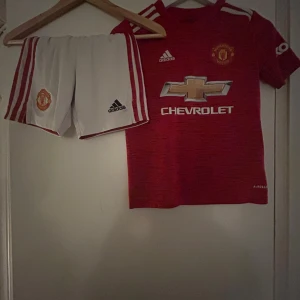 Manchester United fotbollströja Adidas - Säljer en röd Manchester United fotbollströja från Adidas med korta ärmar och vit Chevrolet-logga på bröstet. Tröjan har vita detaljer och klubbmärke. Materialet är lätt och andas, perfekt för match eller träning. Ingår vita shorts med röda ränder och klubbmärke.