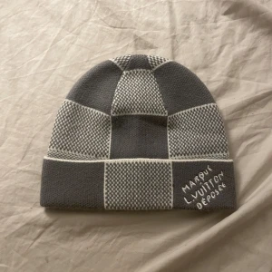 Damier Herutage gråvit rutig mössa - Snygg mössa  i grått och vitt med rutigt mönster. Broderad text 'Marque L. Vuitton Deposee' framtill. Tillverkad i 60% kashmir och 40% ull för extra mjuk och lyxig känsla. Perfekt accessoar för att lyfta din vinterstil.