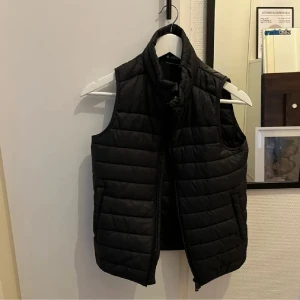 Svart dunväst - Svart dunväst från Everest med quiltad design och hög krage. Västen har dragkedja framtill och två sidofickor. Perfekt att slänga över en hoodie för extra värme under kyliga dagar.