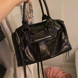 Svart handväska med dragkedja och remmar - Cool svart handväska i skinn med snygga detaljer som dragkedja framtill och långa dekorativa remmar. Väskan har både handtag och axelrem, så du kan bära den precis som du vill. Mått: 30x20x10 cm. Perfekt storlek för dina viktigaste grejer.