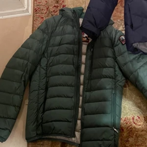 Grön dunjacka med huva från Parajumpers - Säljer en mörkgrön dunjacka från Parajumpers med huva och snygga svarta dragkedjor. Jackan har quiltad design och patch på ärmen. Perfekt för kalla dagar och har en clean look som funkar till det mesta.