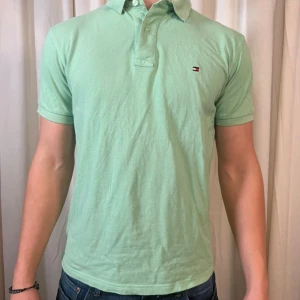 Mintgrön piké från Tommy Hilfiger - Fräsch mintgrön pikétröja från Tommy Hilfiger med klassisk krage och korta ärmar. Tröjan har en liten broderad logga på bröstet och knappar vid halsen. Tillverkad i mjuk bomull som känns skön mot huden. Perfekt för dig som gillar stilrena och enkla plagg.