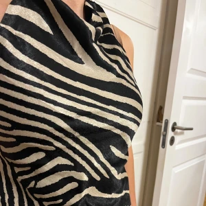 Zebramönstrad halter topp i satin - Säljer en snygg halter topp med zebramönster i svart och beige. Toppen är axelbandslös och har en glansig satinfinish som ger en lyxig känsla. Passformen är tight och den har en cool omlott-detalj framtill. Perfekt för dig som vill sticka ut med ett djuriskt mönster.