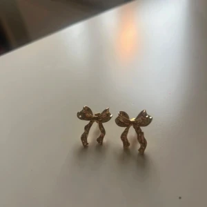 Guldiga örhängen med rosettform - Supersöta örhängen i guld med rosettformad design. Perfekta för dig som gillar detaljer och vill ha något unikt. De är små och diskreta men ändå ett statement med sin glansiga finish och fina form.