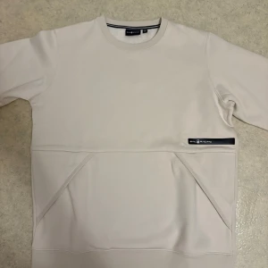 Beige sweatshirt från Sail Racing - Snygg beige sweatshirt från Sail Racing med stilren design och diskret logga på bröstet. Tröjan har rund hals, långa ärmar och praktisk ficka framtill. Perfekt för dig som gillar minimalistisk stil och vill ha något bekvämt och enkelt.