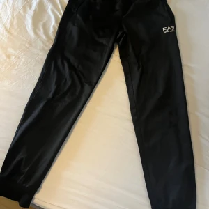 Svarta mjukisbyxor EA7 Emporio Armani - Svarta mjukisbyxor från EA7 Emporio Armani med logga på vänster lår. Byxorna har elastisk midja med snörning och ribbade muddar vid benslut. Perfekta för chill eller träning, riktigt snygga och sköna.