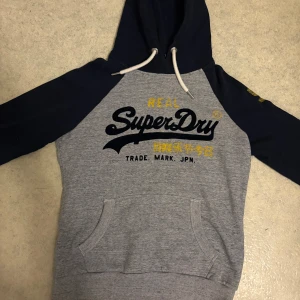 Grå och marinblå Superdry hoodie - Snygg hoodie från Superdry i grått med marinblå ärmar och huva. Stor tryckt logga på bröstet och siffran 54 i gult på ena ärmen. Mjuk insida, känguruficka och vita snören vid huvan. Perfekt för chill dagar och streetstyle.