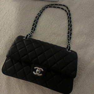Svart quiltad axelväska  - Axelväska i svart skinn med klassiskt quiltat mönster och silverfärgad kedja. Väskan har ikoniskt spänne framtill och vinrött foder på insidan. Perfekt storlek för det viktigaste och en riktig statement piece.