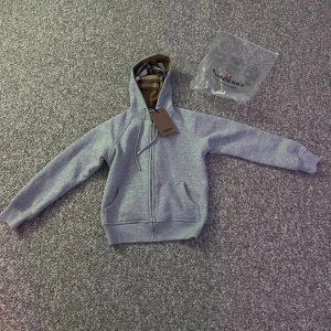 Grå Burberry hoodie med dragkedja - Säljer en grå hoodie från Burberry med klassiskt rutigt foder i huvan och broderad logga på bröstet. Tröjan har dragkedja framtill, fickor och är tillverkad i mjuk bomull. Perfekt för dig som vill ha en stilren och lyxig streetwear-look.