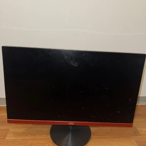 AOC 24-tums LED Monitor - Snygg AOC 24-tums LED-skärm med tunn ram och röd detalj. Skärmen har några synliga fingeravtryck och mindre repor på ytan, men är i gott skick och fungerar utmärkt. Perfekt för både arbete och gaming. Stabil fot och modern design.