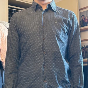 Mörkblå skjorta med småprickigt mönster - Snygg mörkblå skjorta med små vita prickar och klassisk krage. Skjortan har bruna knappar framtill och ett litet broderat emblem på bröstet. Långärmad och i mjukt bomullsmaterial, perfekt för dig som gillar stilrena detaljer. Pris kan diskuteras!