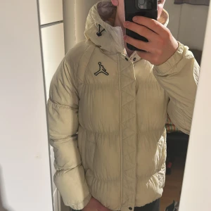 Beige pufferjacka från Jordan - Säljer en fet pufferjacka från Jordan med klassisk logga på bröstet. Jackan har huva med snörning och dragkedja framtill. Fylld för att hålla dig varm under kalla dagar och har en clean, sportig look som passar streetwear-stilen. Det är en xs men den passar mig och jag brukar ha M, 180, 75kg. Om du kollar sista bilden så är det en fläck på baksidan av höger ärm! Det är inget man lägger märke till men kan vara bra att nämna😊