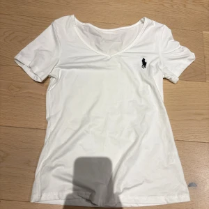 Vit v-ringad t-shirt från Ralph Lauren - Vit t-shirt från Ralph Lauren med v-ringning och korta ärmar. Klassisk design med den ikoniska svarta polospelaren broderad på bröstet. Tillverkad i mjuk bomull som känns skön mot huden. Perfekt för en clean och stilren look.