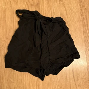 Svarta högmidjade shorts från Monki - Säljer ett par svarta högmidjade shorts från Monki med snygg knytning i midjan. De har en lös passform och är tillverkade i ett lätt tyg som känns skönt mot huden. Perfekta för varma dagar när du vill ha en chill och trendig look.