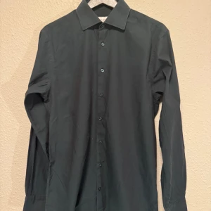 Mörk grön skjorta från John Henric - Stilren mörkgrön skjorta från John Henric med klassisk krage och knappar framtill. Skjortan är långärmad och har en snygg, smal passform som passar perfekt till jeans eller chinos. Tillverkad i mjuk bomull för en skön känsla hela dagen.