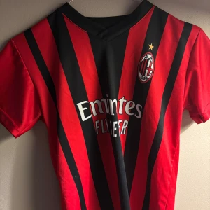 AC Milan matchtröja röd/svart - Säljer en klassisk AC Milan fotbollströja med röda och svarta vertikala ränder, klubbmärke på bröstet och Emirates sponsortryck. Tröjan har korta ärmar och V-ringning. Perfekt för dig som vill visa ditt stöd för Milan på match eller träning.