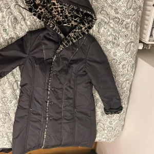 Svart vinterjacka med leopardfoder - Svart lång vinterjacka med huva och snyggt leopardmönstrat foder i pälsimitation. Jackan har knäppning med knappar framtill och markerade sömmar som ger en schysst form. Perfekt för kalla dagar när du vill sticka ut lite extra.