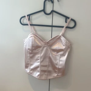 Ljusrosa satin korsett från H&M Divided - Supersöt ljusrosa korsett-topp i satin från H&M Divided. Toppen har smala axelband, markerade kupor och en liten rosett framtill. Snygg croppad passform och glansigt material som ger en lyxig känsla.