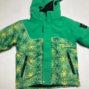 Grön mönstrad vinterjacka Lindberg - Snygg grön vinterjacka från Lindberg med coolt stjärnmönster i blått och gult. Jackan har huva, dragkedja och två fickor med svarta detaljer. Perfekt för kalla dagar och har en schysst passform som funkar för lek och äventyr.