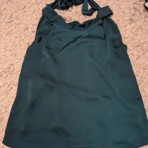 Mörkgrön ärmlös tröja  - Snygg mörkgrön tröja med halterneck och knytning i nacken. Tröjan är ärmlös och har en mjuk, glansig yta som ger en elegant känsla. Perfekt för dig som vill sticka ut med en stilren look. Storleken står inte men gissar på S ☺️