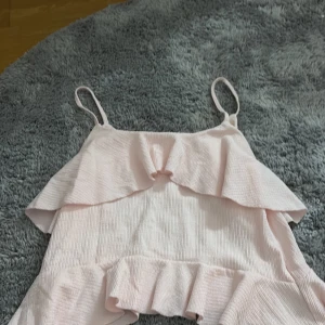 Ljusrosa blusar från Nelly - Supersöt ljusrosa blusar från Nelly i storlek XS. Klänningen har tunna axelband och flera volangdetaljer upptill och nertill. Materialet är lätt och känns som bomull eller polyester, perfekt för varma dagar. Enkel och feminin look som är lätt att styla.