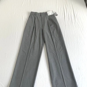Helt nya Uniqlo plisserade vida byxor i grått, storlek S - Helt nya, Snygga grå kostymbyxor från Uniqlo i storlek S. Byxorna har hög midja, vida ben och pressveck framtill. Klassisk look med bälteshällor och bakficka med knapp.