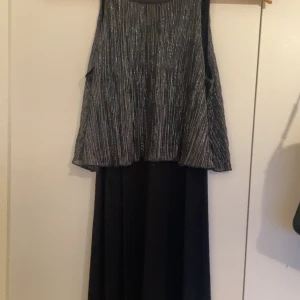Svart långklänning med glittrig topp - Snygg svart långklänning med ärmlös design och glittrig, silverrandig topp. Kjolen är enfärgad svart och faller rakt ner. Perfekt för dig som vill sticka ut med en elegant och modern look.