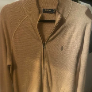 Beige kofta från Polo Ralph Lauren - Säljer en beige stickad kofta från Polo Ralph Lauren med dragkedja framtill och klassisk broderad logga på bröstet. Koftan är i mjuk bomullsblandning och har långärmad design med ribbade muddar och krage. Passar dig som är mellan 178-185