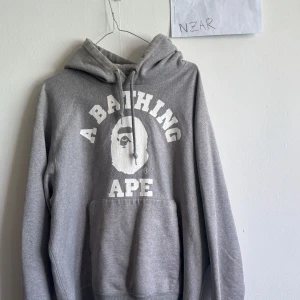 Grå hoodie från A Bathing Ape Bape - Säljer en grå hoodie från A Bathing Ape med stor vit logotyp och apans ansikte tryckt på bröstet. Tröjan har huva med snörning och en klassisk känguruficka framtill. Perfekt för dig som gillar streetwear och vill sticka ut med ett ikoniskt märke.