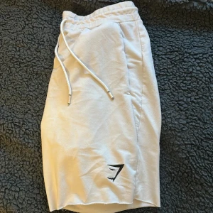 Vita shorts från Gymshark med snörning - Säljer ett par vita shorts från Gymshark med elastisk midja och snörning. De har en avslappnad passform och är tillverkade i mjuk bomull, perfekt för träning eller chill. Gymshark-loggan syns diskret på ena benet. Snygga och enkla att matcha med det mesta.