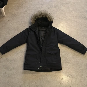 Svart parkajacka från Everest med fuskpäls storlek 158 - Svart parkajacka från Everest med huva och fuskpäls. Jackan har stora fickor framtill, dragkedja och knappar. Perfekt för kalla dagar med sin långa modell och fodrade insida. Snygg och funktionell design som håller dig varm hela vintern.
